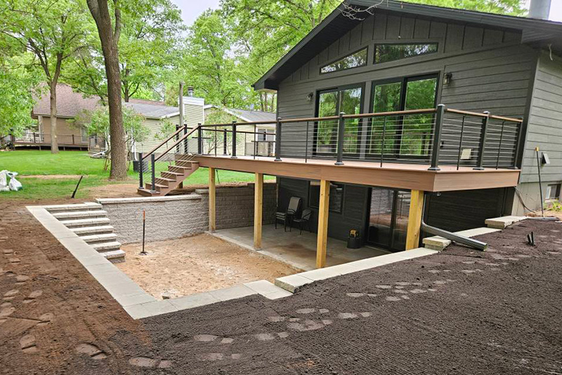 New deck with cable railing, retaining wall, and paver steps under construction in a wooded residential backyard