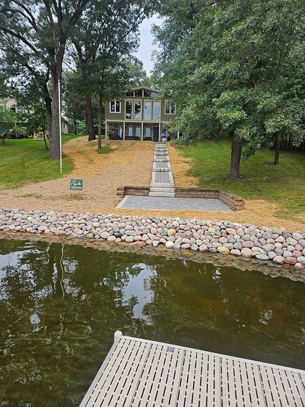 Completed lakefront landscaping with a paver staircase, boulder shoreline, and freshly seeded yard viewed from the dock
