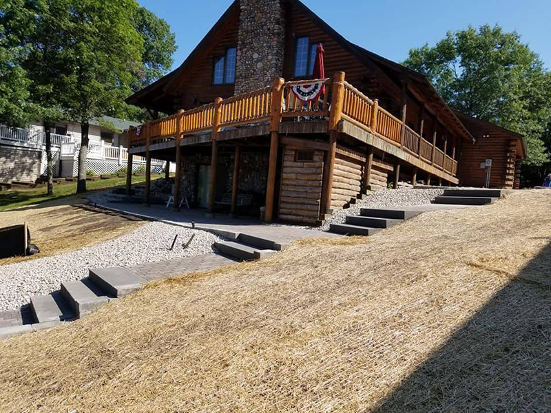 New paver steps and gravel patio area with freshly seeded slope alongside a log cabin with a wraparound deck