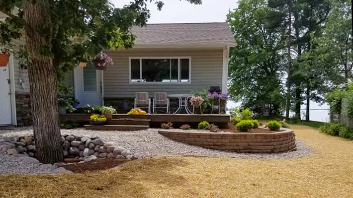 Lakefront cottage with a curved retaining wall, flower-filled deck, gravel drainage area, and freshly seeded yard