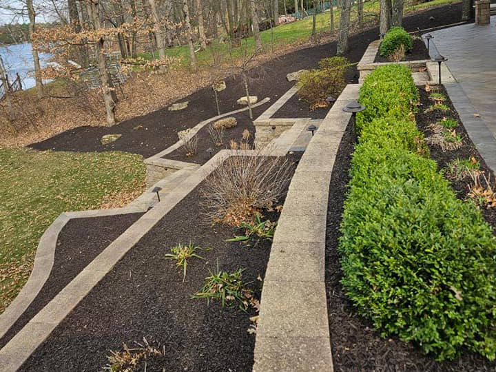 Tiered garden beds with stone edging, boxwood shrubs, landscape lighting, and dark mulch on a sloped lakefront property