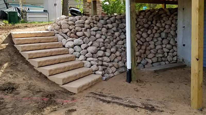 Paver steps and round boulder retaining walls under construction beneath a raised deck addition