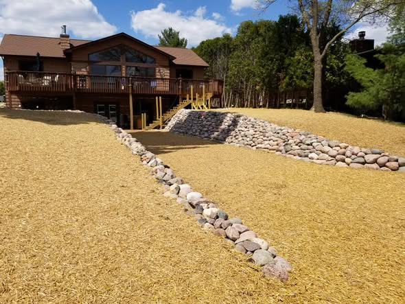 Freshly seeded yard with straw coverage, boulder drainage swales, and paver steps leading to a lakefront cabin deck